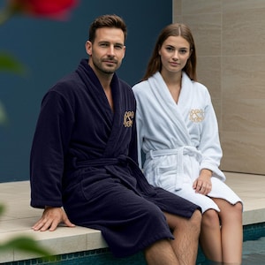 May include: Two bathrobes, one navy blue and one white, with gold embroidered monograms. The robes are worn by two people sitting on a ledge by a pool.
