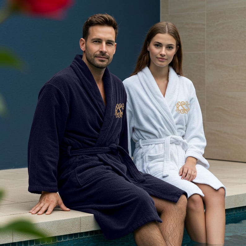 May include: Two bathrobes, one navy blue and one white, with gold embroidered monograms. The robes are worn by two people sitting on a ledge by a pool.