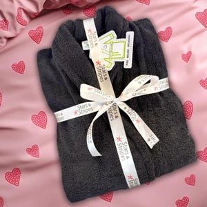 Puede incluir: Una bata gris oscuro atada con una cinta blanca impresa con "Stars & Stripes". La bata se presenta sobre un fondo rosa con patrones de corazones rojos. La bata tiene una etiqueta que dice "Pure Organic Sustainable".