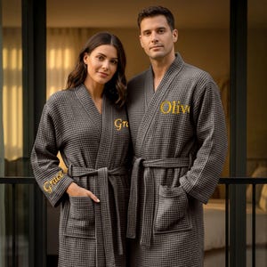 May include: Two people wearing matching grey waffle-weave bathrobes. Each robe has a belt, pockets, and gold embroidered text on the chest and sleeve. The text on one robe says "Grace" and the other says "Olive".