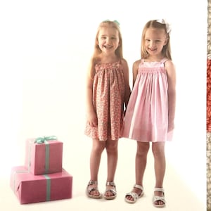May include: Two young girls wearing dresses, one floral and one solid pink, stand side-by-side. They are smiling and wearing sandals. Pink wrapped gift boxes with teal ribbons are in the foreground. The word "KATINA" is at the top.