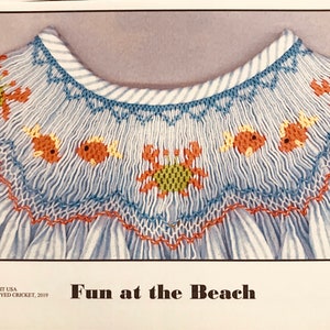 May include: A close-up of a blue and white striped fabric with a white embroidered design of crabs and fish. The fabric is folded over and the embroidery is visible on both sides. The text "Fun at the Beach" is printed below the fabric.