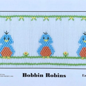 May include: A cross stitch pattern featuring three blue robins with orange breasts, sitting on a green branch with yellow flowers. The pattern is titled "Bobbin Robins" and is described as "Easy to Stitch".
