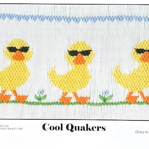 May include: Three yellow ducklings wearing sunglasses, with orange feet, are embroidered on a white fabric with green grass and blue flowers. The text "Cool Quakers (Easy to Smock)" is below the ducks.