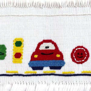 May include: A white fabric with a red car, a traffic light, a green shape, a red circle with a smiley face, and a yellow key. The fabric has a fringe border.