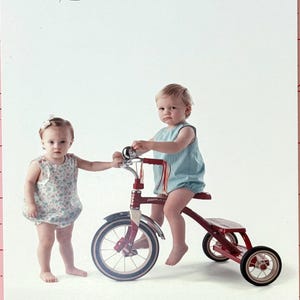 May include: Two children, one girl and one boy, are playing with a red tricycle. The girl is wearing a white floral dress and the boy is wearing a light blue sleeveless shirt. The tricycle has a white seat and a white handlebar with a red frame.