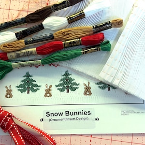May include: A craft project featuring embroidery floss in brown, white, gold, red, and green. The project includes a printed design of Christmas trees and bunnies, with the text "Snow Bunnies". A red and white ribbon is also included.
