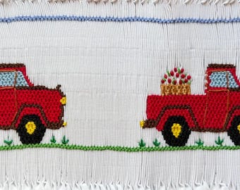 Puede incluir: Tela blanca bordada con dos camionetas rojas que transportan cestas de flores. Los camiones tienen ruedas negras y faros amarillos. Hierba verde y una franja azul en la parte superior e inferior.