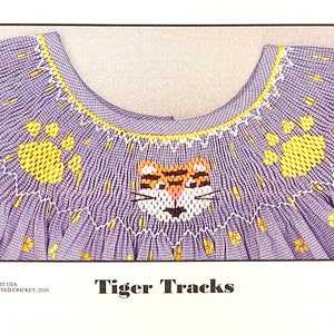 May include: A purple and white gingham fabric with yellow embroidered paw prints and a tiger face. The text "Tiger Tracks" is printed below the image. Copyright USA © CROSS-EYED CRICKET, 2020. Plate #4333.