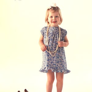 May include: A young child wearing a blue floral dress and a pearl necklace. The dress has a ruffled hem. Gold high heels are on the floor. The text "MIA" and "sizes 6mo-6" are visible.