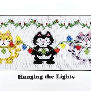 May include: Three cute cats, one yellow, one black, and one gray, are hanging colorful Christmas lights on a green garland. The text "Hanging the Lights" is below the cats.