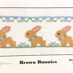 May include: A cross-stitch pattern featuring three brown bunnies with pink ears and white tails, each wearing a green and blue collar. The bunnies are set against a white background with blue and green floral accents. The text "Brown Bunnies" is displayed.
