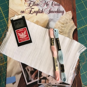 May include: A book titled "Ellen Me Carn on English Smocking" with a white fabric, two spools of embroidery floss in pink and light blue, and a package of needles.