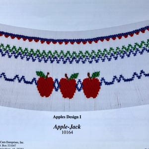 May include: A white fabric with a blue, green, and red embroidered design of three apples with leaves. The fabric is labeled "Apples Design I Apple-Jack 10164".