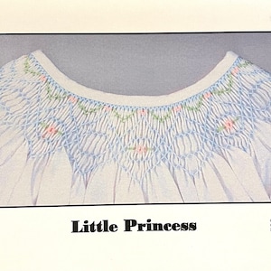 May include: White baby dress with a smocked bodice featuring light blue and pink floral embroidery. The dress has a round neckline and pleated skirt. The text "Little Princess" is displayed at the bottom.