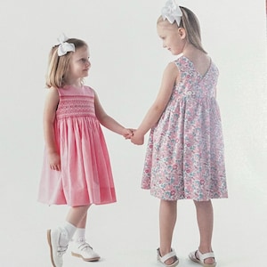 May include: A sewing pattern envelope titled MARY DE by Children's Corner Patterns for sizes 6mo-3. The cover features two sleeveless dresses: one solid pink with a smocked bodice and one floral print with a V-back, both worn with white hair bows.