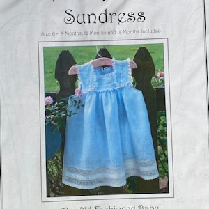 May include: A sewing pattern for a French Acadian sundress for babies, sizes 6-9 months, 12 months, and 18 months. The pattern is by Jeannie Downs Baumeister and features a light blue dress with white lace trim.