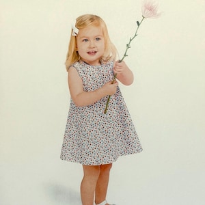May include: A young child wearing a sleeveless floral print dress and white sandals, holding a pink flower. The dress features a pattern of small red, blue, and white flowers on a light background. A white bow is in the child's hair.