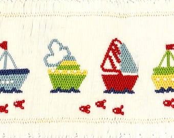 Puede incluir: Un panel de tela blanca con una fila horizontal de cuatro barcos bordados en azul, verde, rojo y amarillo. Cada barco tiene una pequeña bandera roja o detalle de humo, y pequeños peces rojos están bordados debajo de cada embarcación.