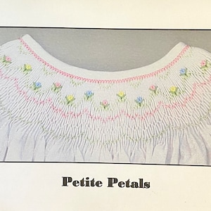 May include: White smocked garment with a rounded neckline, featuring delicate embroidery in pink, yellow, and blue floral patterns. The text "Petite Petals" is displayed below the garment.