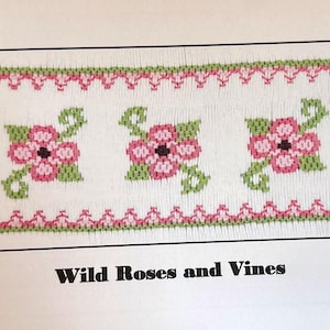 May include: A cross-stitch pattern featuring three pink flowers with black centers and green leaves, framed by pink and green decorative borders. The text "Wild Roses and Vines" is displayed below the design.