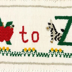 May include: A white fabric panel featuring cross-stitch embroidery with the text A to Z in green and red. The letter A is paired with a red apple, and the letter Z is paired with a black and white zebra. A row of yellow flowers with green stems runs below.