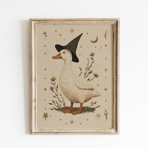 Pode incluir: Uma impressão de arte emoldurada apresentando um pato branco usando um chapéu preto pontudo. O pato está sobre um pequeno pedaço de grama verde cercado por ilustrações botânicas, pequenas estrelas e uma lua crescente em fundo bege.