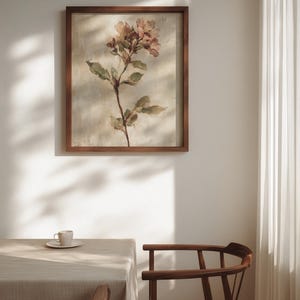 May include: A framed floral print featuring a watercolor-style depiction of a flowering stem with pink and green hues. The artwork is set in a dark brown wooden frame, hanging on a white wall. A table and chair are in the foreground.