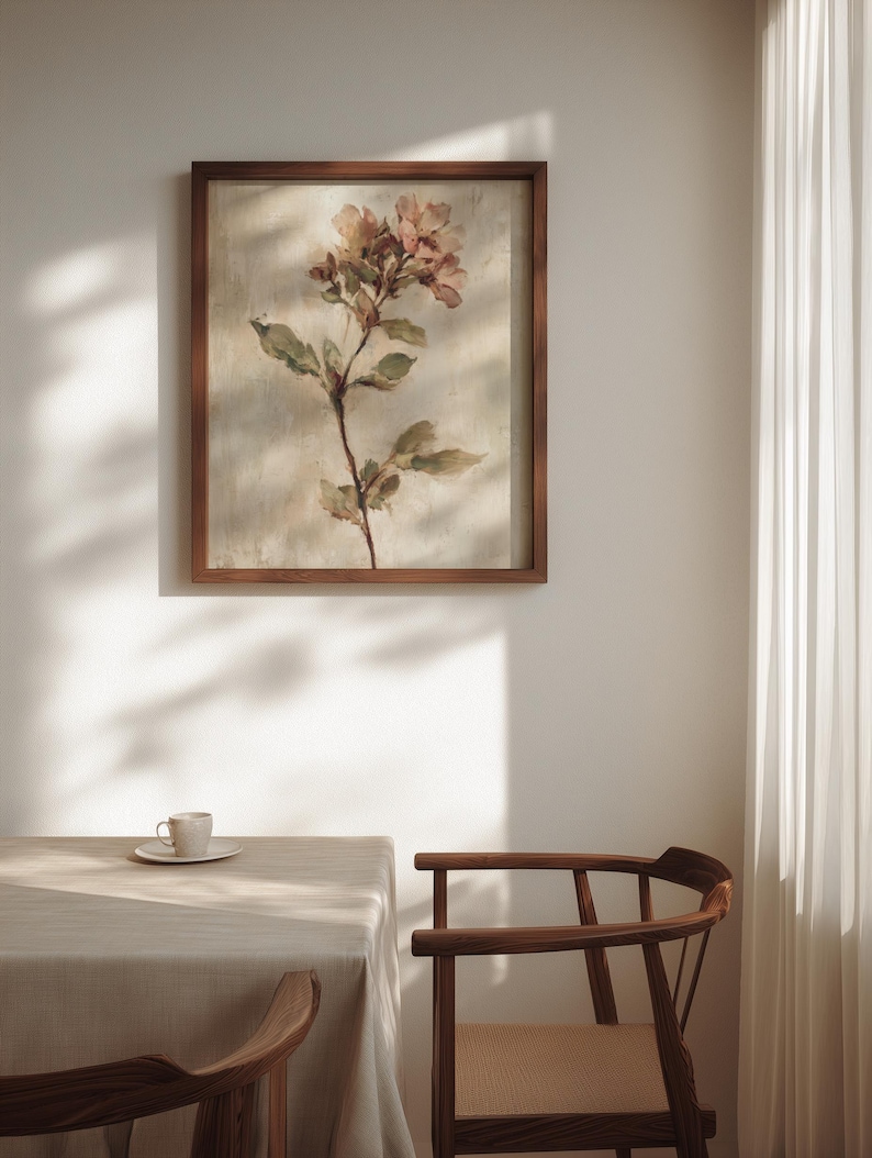 May include: A framed floral print featuring a watercolor-style depiction of a flowering stem with pink and green hues. The artwork is set in a dark brown wooden frame, hanging on a white wall. A table and chair are in the foreground.