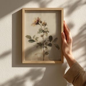 Puede incluir: Una impresi&oacute;n bot&aacute;nica enmarcada con una ilustraci&oacute;n en acuarela de una planta floreciente con delicados p&eacute;talos rosas y crema, hojas verdes y un marco de madera marr&oacute;n claro. La obra de arte est&aacute; sostenida por una persona.