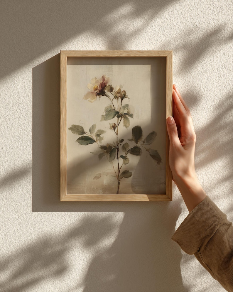 Puede incluir: Una impresi&oacute;n bot&aacute;nica enmarcada con una ilustraci&oacute;n en acuarela de una planta floreciente con delicados p&eacute;talos rosas y crema, hojas verdes y un marco de madera marr&oacute;n claro. La obra de arte est&aacute; sostenida por una persona.