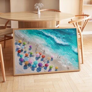 May include: A framed art print showing a beach scene with colourful umbrellas and people. The artwork features a vibrant turquoise ocean meeting a sandy shore, with the umbrellas in shades of red, pink, yellow, and blue. The frame is made of light-coloured wood.