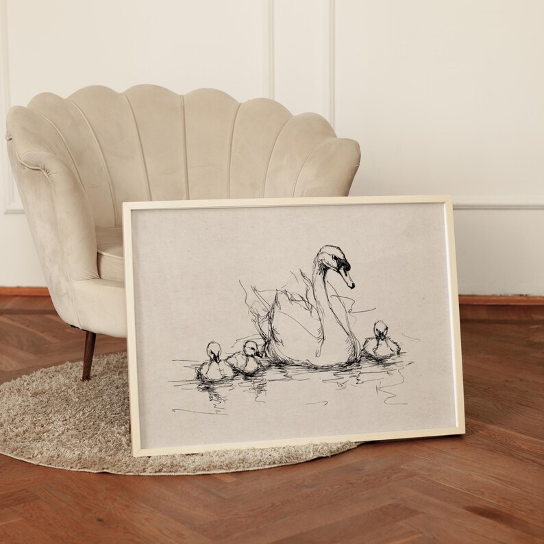 May include: Framed black and white sketch of a swan with three cygnets swimming in water. The artwork is in a light-colored wooden frame, resting on a rug and hardwood floor. The background includes a beige armchair.