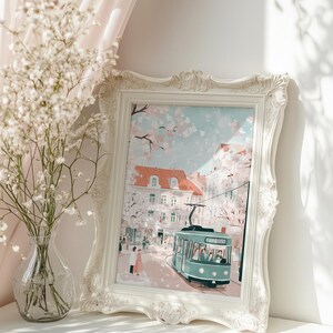 May include: A white framed print of a city scene with a green tram in the foreground. The scene is set in a European city with buildings and trees. The sky is blue with white clouds. The print is on a white shelf with a vase of white flowers in the foreground.