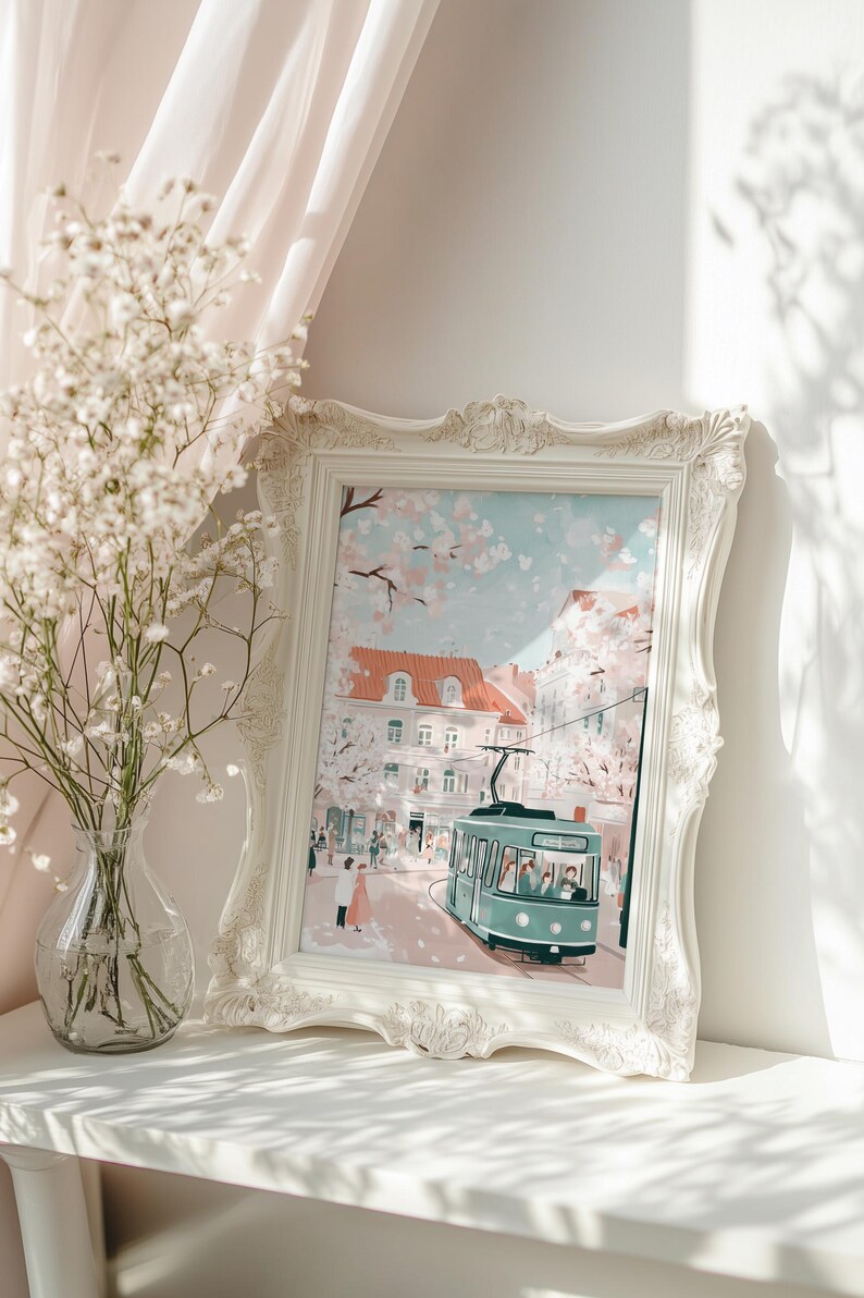 May include: A white framed print of a city scene with a green tram in the foreground. The scene is set in a European city with buildings and trees. The sky is blue with white clouds. The print is on a white shelf with a vase of white flowers in the foreground.