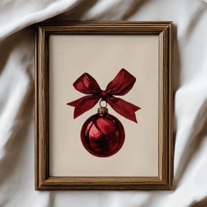 May include: A framed print featuring a red Christmas ornament with a matching bow. The ornament is a deep, glossy red, and the bow is tied at the top. The artwork is set against a cream background and framed in a wooden frame.