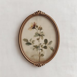 May include: An oval framed floral print in an antique style. The artwork depicts a botanical illustration of a flowering plant with green leaves and a pale yellow and pink flower. The frame is gold-coloured.