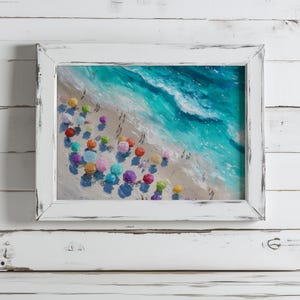 May include: A framed painting of a beach scene in a white, distressed frame. The artwork shows a sandy beach with colourful umbrellas and turquoise waves. People are scattered along the shore, enjoying the sunny day.