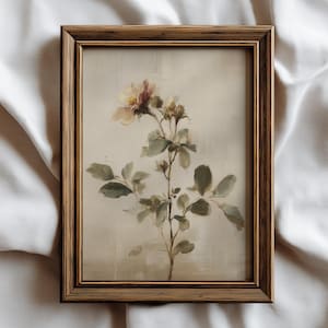 Puede incluir: Una impresi&oacute;n bot&aacute;nica enmarcada que presenta un delicado arreglo floral. La obra de arte muestra una flor de color claro con tonos rosados y amarillos, acompa&ntilde;ada de hojas y tallos verdes. El marco es de un tono c&aacute;lido de madera natural.