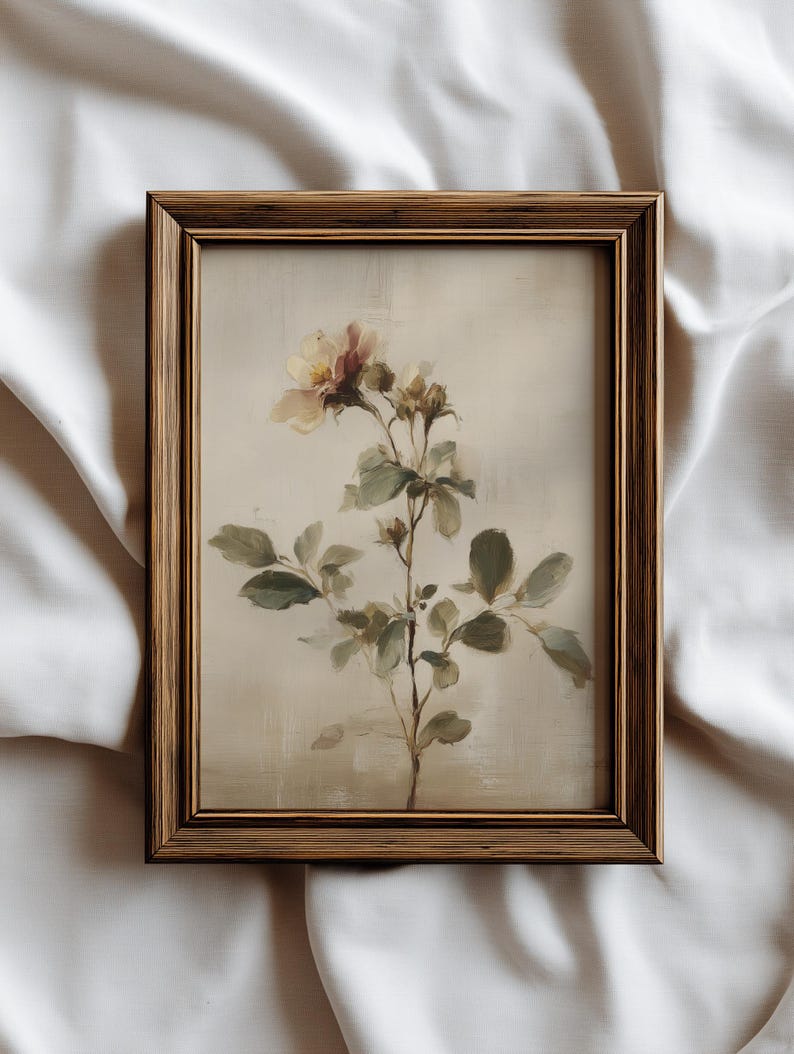 Puede incluir: Una impresi&oacute;n bot&aacute;nica enmarcada que presenta un delicado arreglo floral. La obra de arte muestra una flor de color claro con tonos rosados y amarillos, acompa&ntilde;ada de hojas y tallos verdes. El marco es de un tono c&aacute;lido de madera natural.
