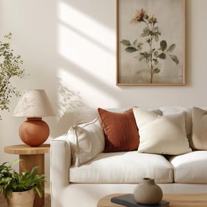 Puede incluir: Un sal&oacute;n con un sof&aacute; blanco y cojines crema y &oacute;xido. Una mesa auxiliar de madera sostiene una l&aacute;mpara de terracota y una planta en maceta. Una impresi&oacute;n bot&aacute;nica enmarcada cuelga de la pared y una peque&ntilde;a mesa de centro de madera est&aacute; frente al sof&aacute;.