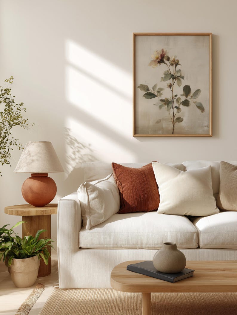 Puede incluir: Un sal&oacute;n con un sof&aacute; blanco y cojines crema y &oacute;xido. Una mesa auxiliar de madera sostiene una l&aacute;mpara de terracota y una planta en maceta. Una impresi&oacute;n bot&aacute;nica enmarcada cuelga de la pared y una peque&ntilde;a mesa de centro de madera est&aacute; frente al sof&aacute;.