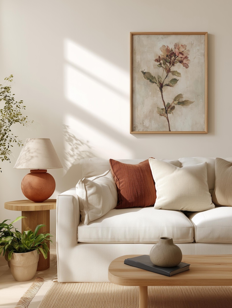 May include: A living room scene featuring a white sofa with rust and cream-colored throw pillows. A framed floral print hangs on the wall. A terracotta lamp sits on a wooden side table, and a small potted plant is nearby.