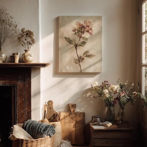 May include: A cozy interior scene featuring a floral painting, a woven basket with a blanket, and wooden cutting boards. Dried flowers in vases and a small wooden crate with a book add to the rustic aesthetic. The room is bathed in warm sunlight.