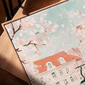 May include: A framed painting of a city street scene with a pink and white cherry blossom tree in the foreground. The buildings have red tile roofs and are painted in soft pastel colors.