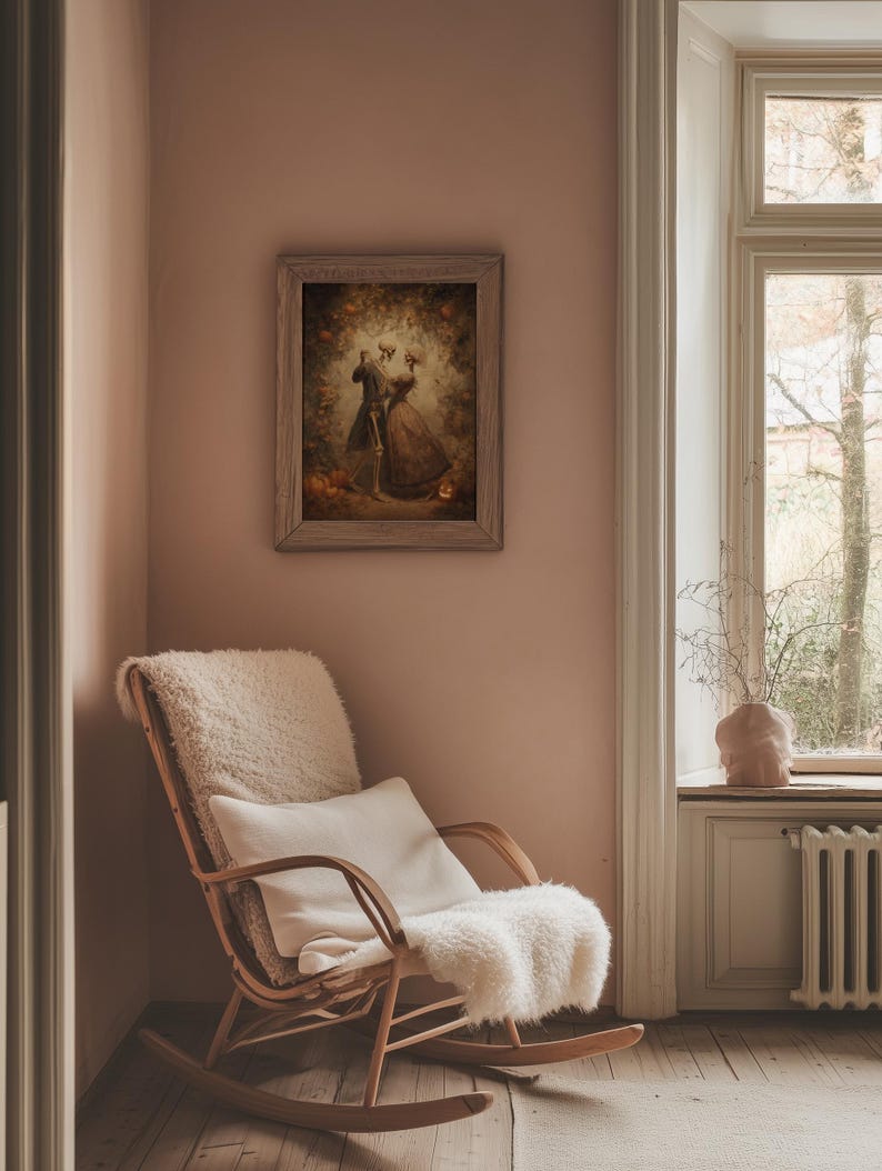 Op de afbeelding: Een ingelijst kunstwerk toont een skeletpaar dat danst in een romantische setting, met herfstbladeren en pompoenen. Het kunstwerk hangt aan een roze muur boven een houten schommelstoel met een wit kussen en een fluffy plaid.