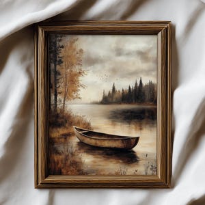 Op de afbeelding: Een ingelijst landschapsschilderij dat een serene meerzicht toont. Een houten boot ligt aan de waterkant, omringd door bomen en gebladerte in herfstkleuren. De lucht is bewolkt en het kunstwerk zit in een houten lijst.
