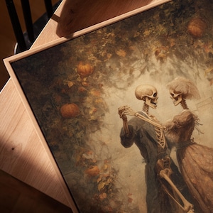Op de afbeelding: Een ingelijste print met een vintage illustratie van twee dansende skeletten. Het kunstwerk heeft een warm kleurenpalet met pompoenen en bladeren op de achtergrond. De skeletten zijn gekleed in kleding uit die tijd.