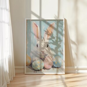 May include: A white rabbit with pink ears sits surrounded by pastel colored Easter eggs. The rabbit is looking at the viewer with a curious expression. The background is a light blue with a shadow of a plant.