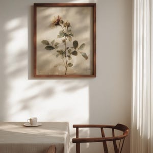 Puede incluir: Una impresi&oacute;n bot&aacute;nica enmarcada con una rama de rosa con hojas verdes y flores rosas y amarillas. La obra de arte est&aacute; en un marco de madera oscura, colgada en una pared blanca. Una mesa con un mantel neutro y una taza y platillo est&aacute;n en primer plano.